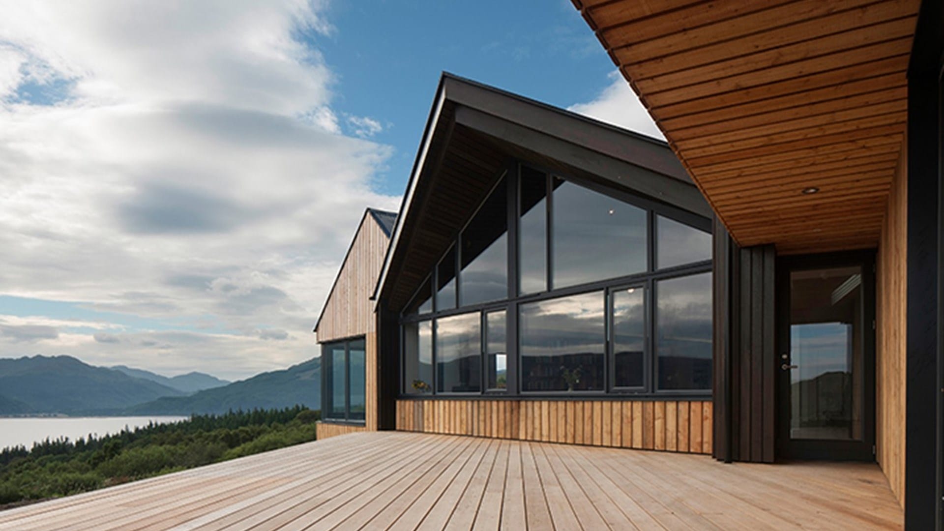 Modern wooden house with large glass windows and a spacious deck, overlooking a scenic landscape with mountains and water under a partly cloudy sky.