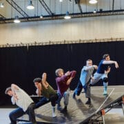 Five dancers perform on an angled platform indoors, each striking expressive, synchronized poses under overhead lights.