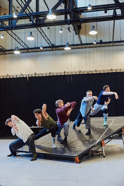 Five dancers perform on an angled platform indoors, each striking expressive, synchronized poses under overhead lights.
