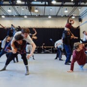 A group of dancers rehearses in a studio, performing energetic lifts, jumps, and poses, with some seated or leaning on the floor.