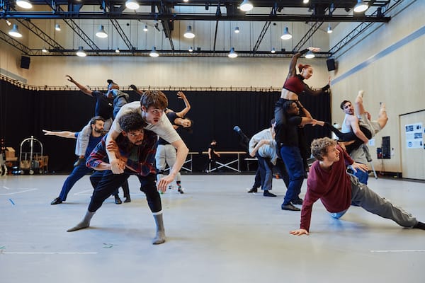 A group of dancers rehearses in a studio, performing energetic lifts, jumps, and poses, with some seated or leaning on the floor.