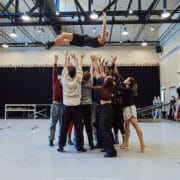 A group of dancers lifts a person high above their heads in a brightly lit studio, while another dancer stands to the side watching.