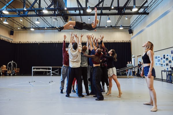 A group of dancers lifts a person high above their heads in a brightly lit studio, while another dancer stands to the side watching.