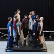 A group of men stands in two facing rows on a raised platform in a rehearsal space with a black curtain backdrop.