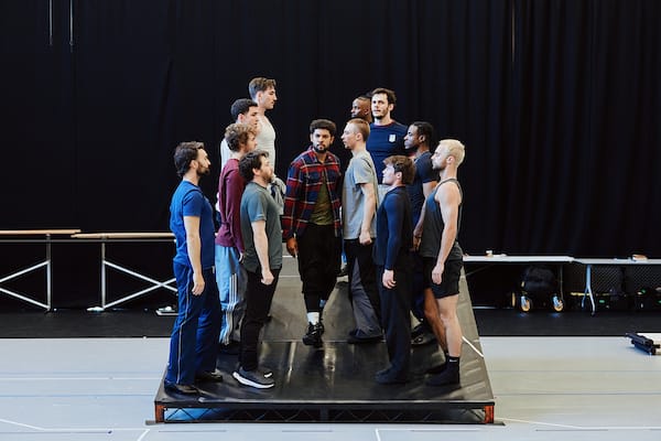 A group of men stands in two facing rows on a raised platform in a rehearsal space with a black curtain backdrop.