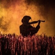 Silhouette of a person playing the violin in a field, with tall grass in the foreground and an orange smoky background—evoking the spirit of Fiddler on the Roof, the beloved Broadway musical.