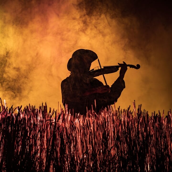 Silhouette of a person playing the violin in a field, with tall grass in the foreground and an orange smoky background—evoking the spirit of Fiddler on the Roof, the beloved Broadway musical.