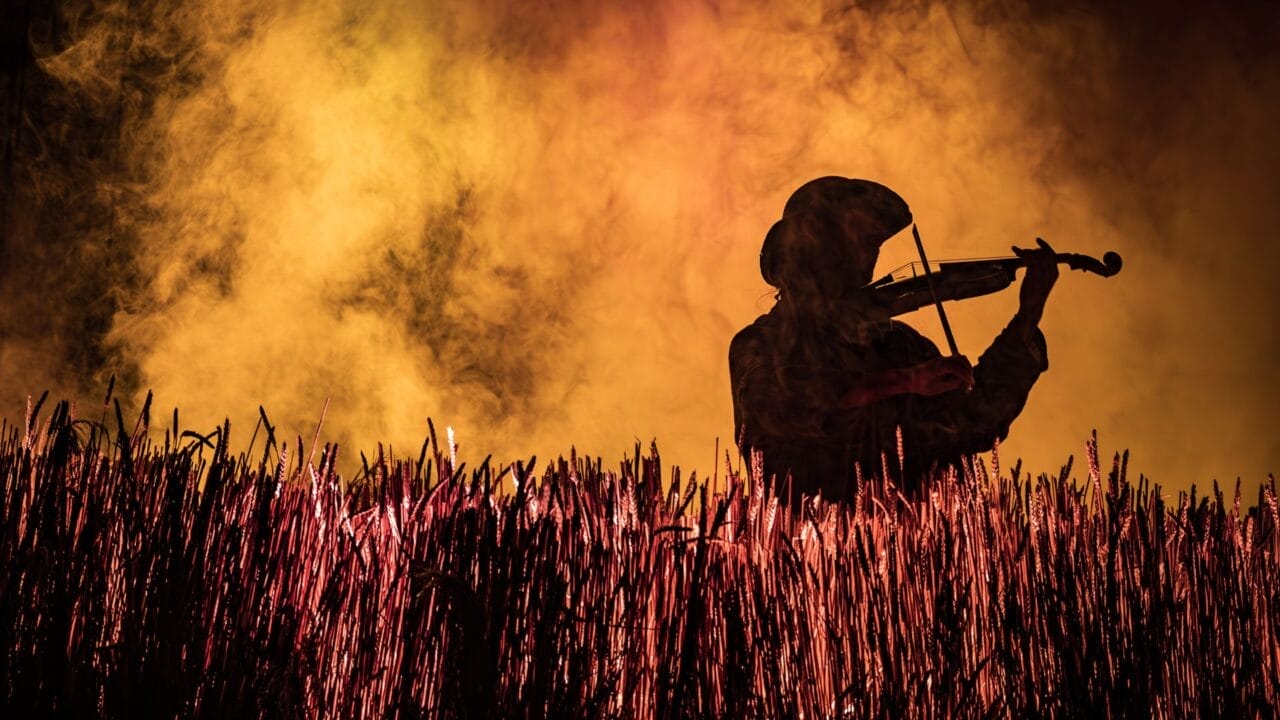 Silhouette of a person wearing a hat and playing a violin, standing in tall grass with yellow and orange mist in the background, evoking the iconic imagery of Fiddler on the Roof, the beloved Broadway musical.