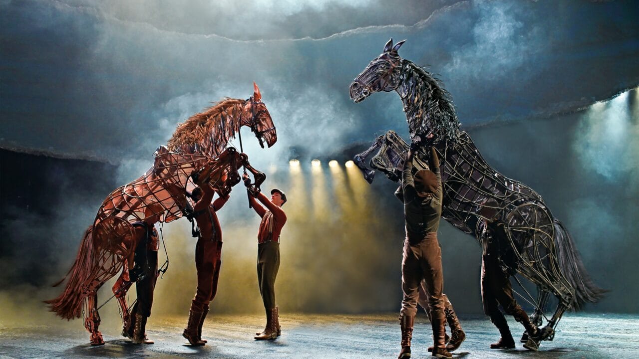 Performers control large, lifelike horse puppets on a misty stage with dramatic lighting and a smoky backdrop.