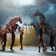 Performers control large, lifelike horse puppets on a misty stage with dramatic lighting and a smoky backdrop.