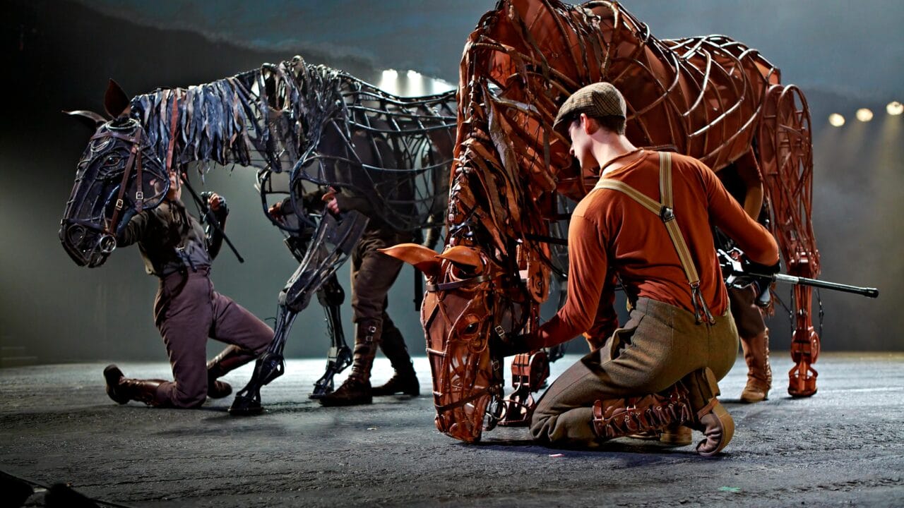 Actors manipulate large, detailed horse puppets on stage during a theatrical performance, with dramatic lighting highlighting the scene.