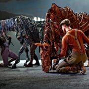 Actors manipulate large, detailed horse puppets on stage during a theatrical performance, with dramatic lighting highlighting the scene.