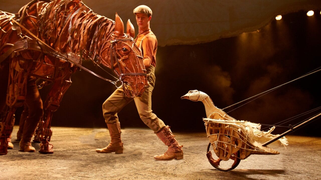 A performer on stage handles a large horse puppet while a goose puppet on wheels, controlled by rods, is positioned nearby under stage lighting.
