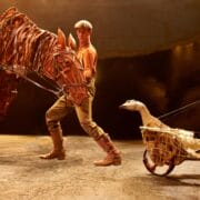 A performer on stage handles a large horse puppet while a goose puppet on wheels, controlled by rods, is positioned nearby under stage lighting.