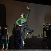 A performer in mid-jump on stage, dressed in period costume, evokes Hans Christian Andersen’s fairy tale, with other actors and a flower cart in the background.