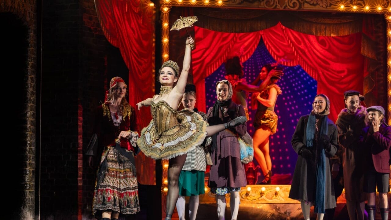 A performer in a gold costume dances on stage with one leg raised, holding a fan, as the Snow Queen and others in period clothing watch. A red curtain and another dancer complete the enchanting scene in the background.