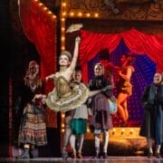 A performer in a gold costume dances on stage with one leg raised, holding a fan, as the Snow Queen and others in period clothing watch. A red curtain and another dancer complete the enchanting scene in the background.