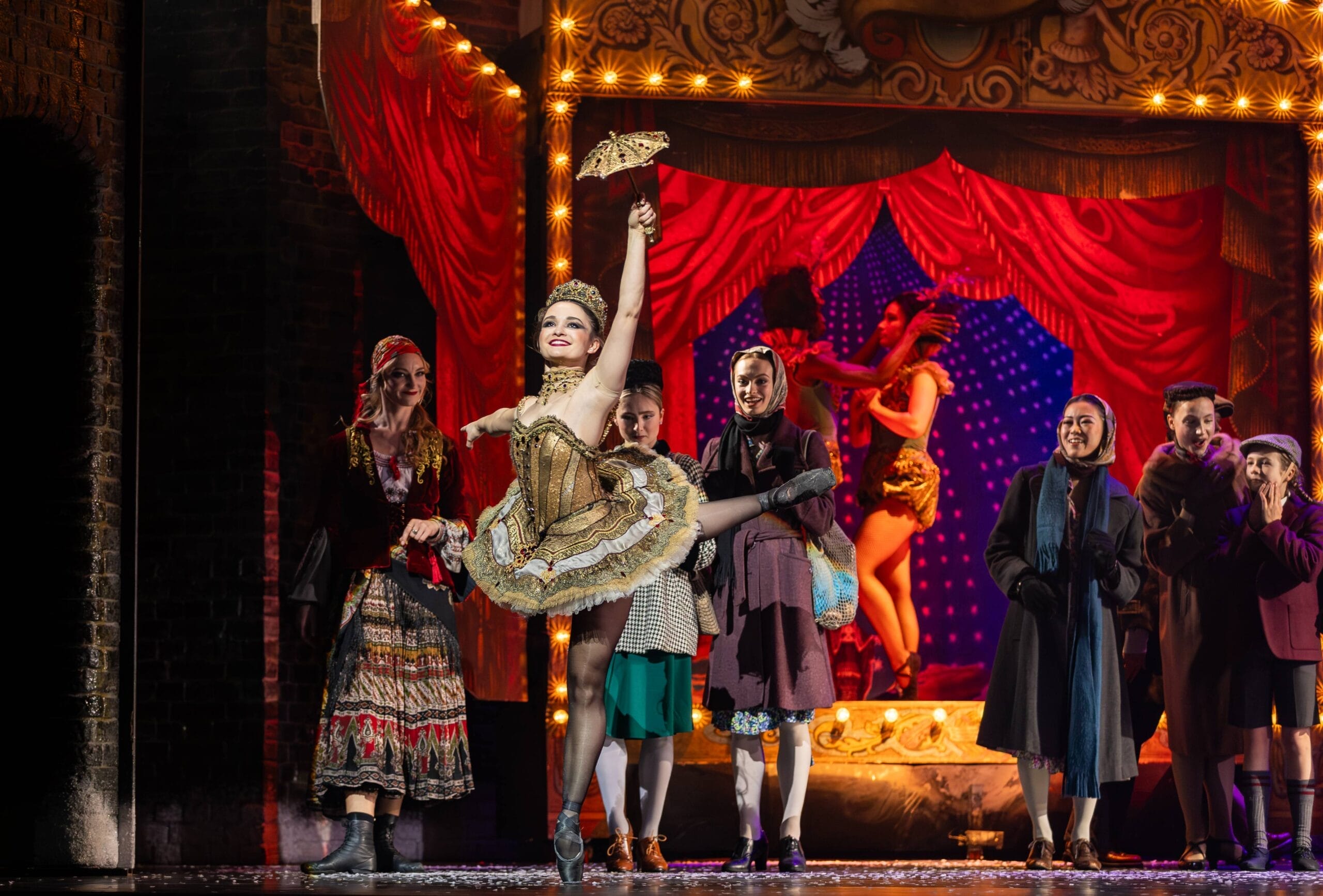A performer in a gold costume dances on stage with one leg raised, holding a fan, as the Snow Queen and others in period clothing watch. A red curtain and another dancer complete the enchanting scene in the background.