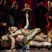 Two circus performers pose dramatically on stage, evoking the magic of The Snow Queen, with one holding a prop umbrella and leaning over the other, while a dressed audience looks on in the background.