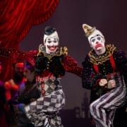 Two clowns in colorful, harlequin-patterned costumes and makeup perform on stage during a lively scene from The Snow Queen, with other actors and a red curtain in the background.