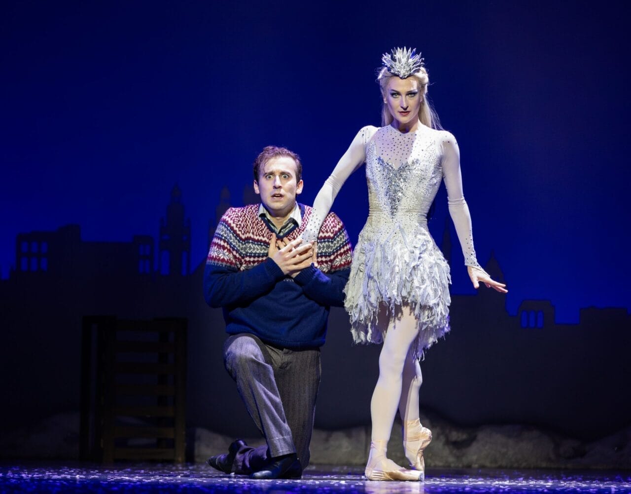 A man kneels on stage clutching his chest beside the Snow Queen, a ballerina in a white, icy costume and crown; a blue backdrop with a city silhouette is behind them.