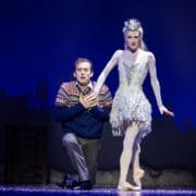 A man kneels on stage clutching his chest beside the Snow Queen, a ballerina in a white, icy costume and crown; a blue backdrop with a city silhouette is behind them.