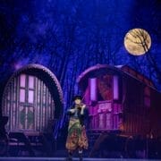 A performer plays violin on stage as the Snow Queen, set before two colorful wooden wagons, with a backdrop of trees and a full moon.