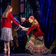 Two women in colorful costumes perform on stage; one stands while the other leans forward with dramatic hand gestures, bringing The Snow Queen to life against a theatrical backdrop.