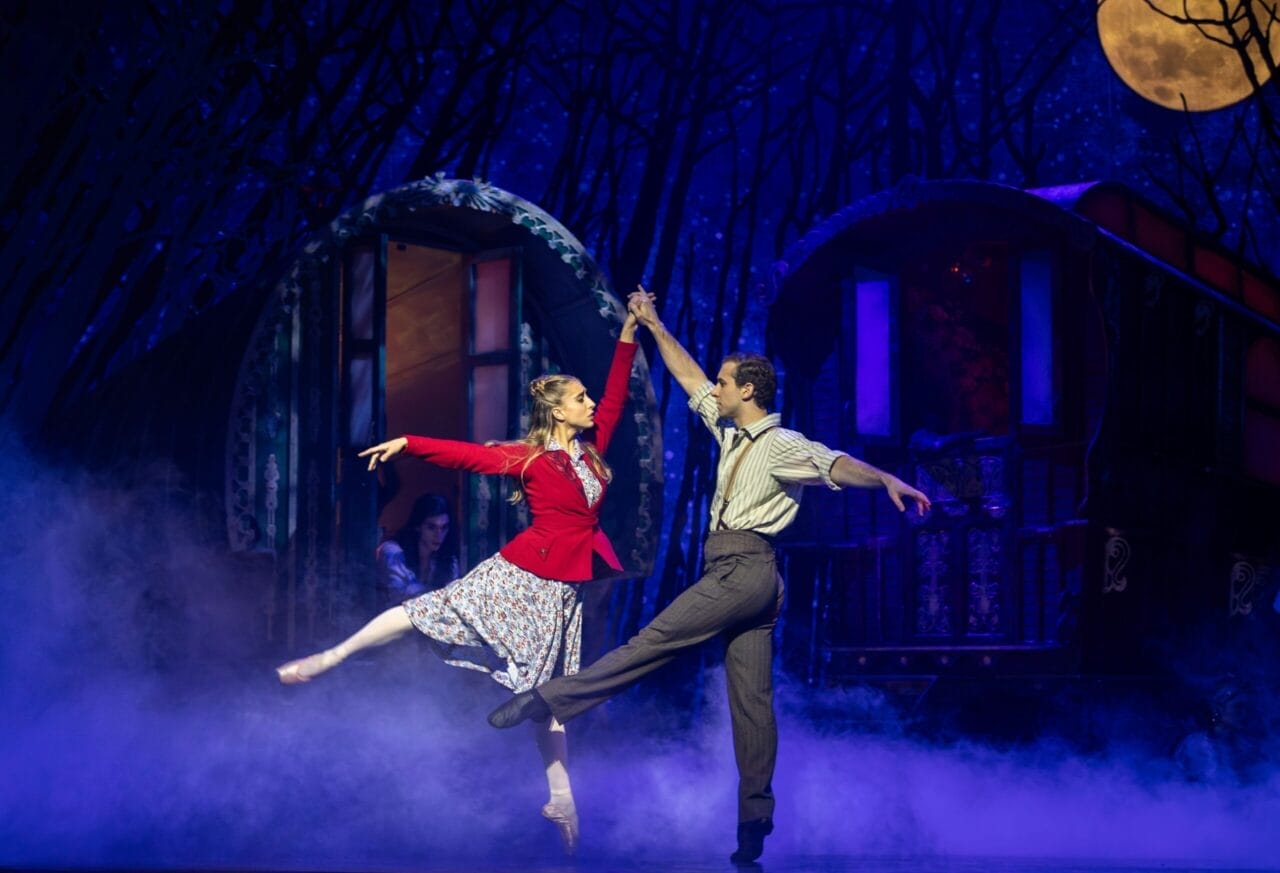 Two dancers perform a ballet duet on a misty stage, with painted caravan wagons and a large moon in the background, evoking the enchanting world of The Snow Queen.