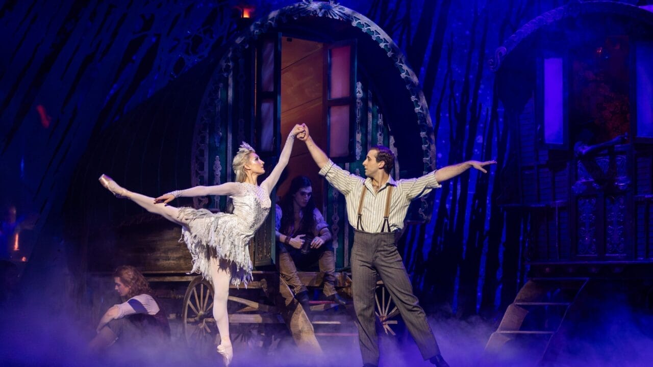 A ballerina and a male dancer perform a duet on stage in front of caravan wagons, evoking a fairy tale atmosphere with mist and two seated figures in the background.