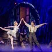 A ballerina and a male dancer perform a duet on stage in front of caravan wagons, evoking a fairy tale atmosphere with mist and two seated figures in the background.