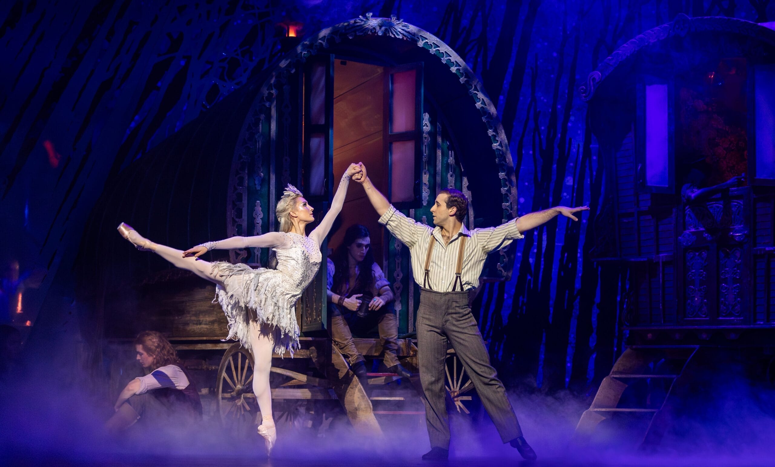 A ballerina and a male dancer perform a duet on stage in front of caravan wagons, evoking a fairy tale atmosphere with mist and two seated figures in the background.