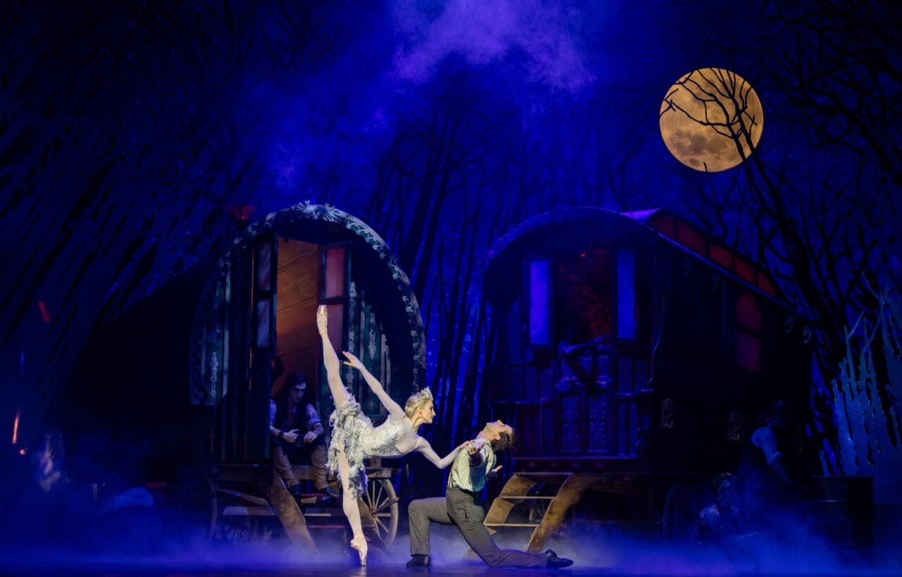 A ballerina and dancer perform a dramatic pose on stage, evoking the magic of Hans Christian Andersen’s Snow Queen, with two wagon-like structures and a full moon in a misty, blue-lit forest background.