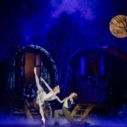 A ballerina and dancer perform a dramatic pose on stage, evoking the magic of Hans Christian Andersen’s Snow Queen, with two wagon-like structures and a full moon in a misty, blue-lit forest background.