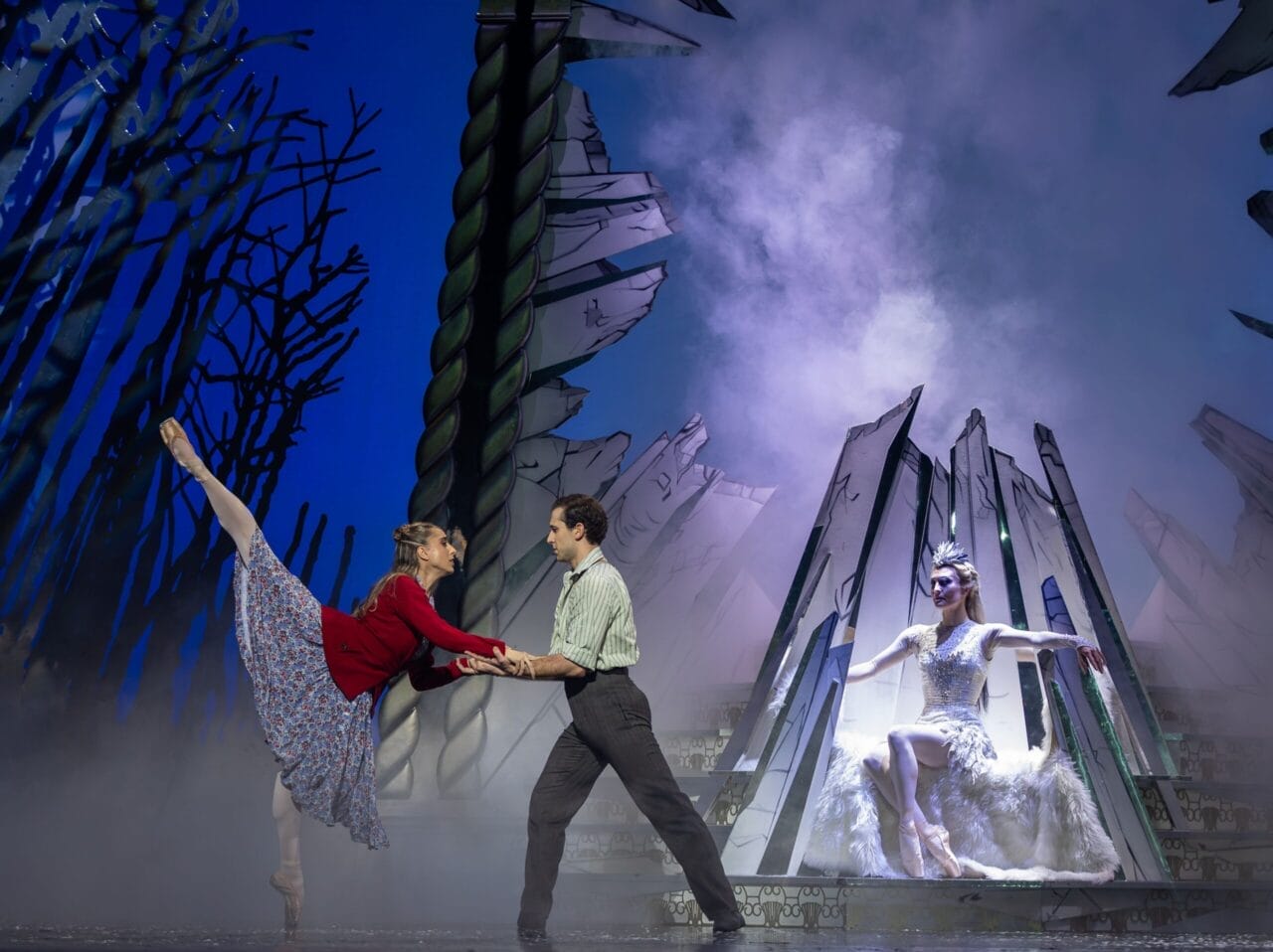 Two dancers perform a lift on stage with a forest backdrop, while The Snow Queen, dressed in white, sits regally on a throne-like structure in the background.