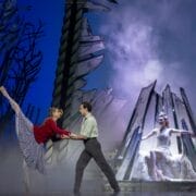 Two dancers perform a lift on stage with a forest backdrop, while The Snow Queen, dressed in white, sits regally on a throne-like structure in the background.