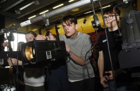 Several people adjust stage lighting equipment in an indoor setting, focusing intently on their tasks under fluorescent overhead lights.
