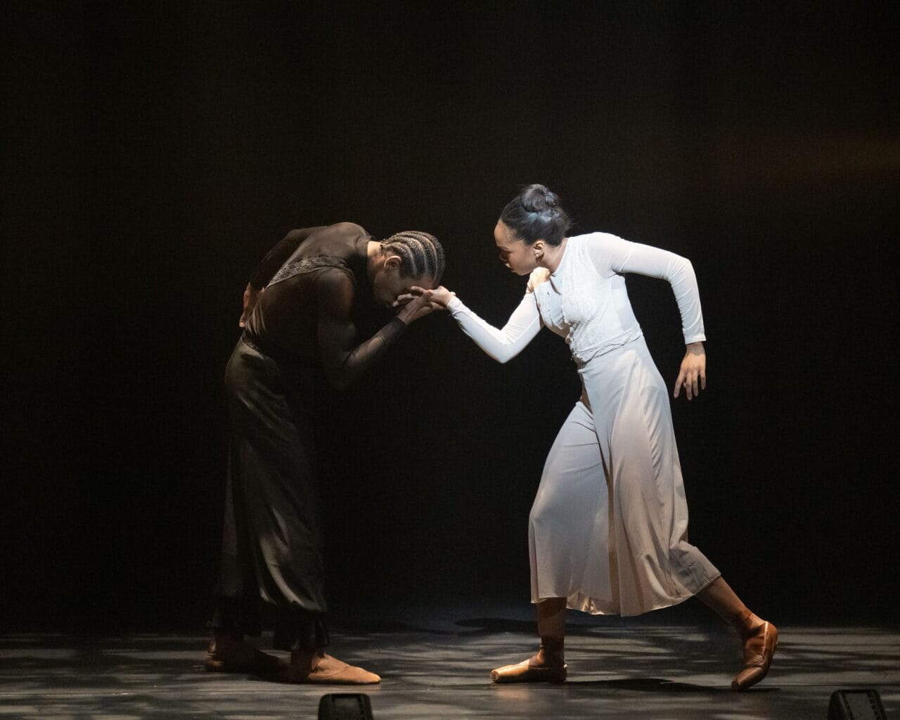 Two dancers perform on stage; one in black bends forward while the other in white extends an arm, their hands touching, both wearing dance shoes and dramatic lighting.