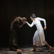 Two dancers perform on stage; one in black bends forward while the other in white extends an arm, their hands touching, both wearing dance shoes and dramatic lighting.