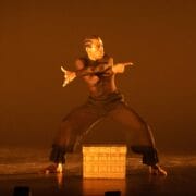 A dancer in black clothing performs a dramatic pose on stage in front of a wooden crate, against an orange-lit background.