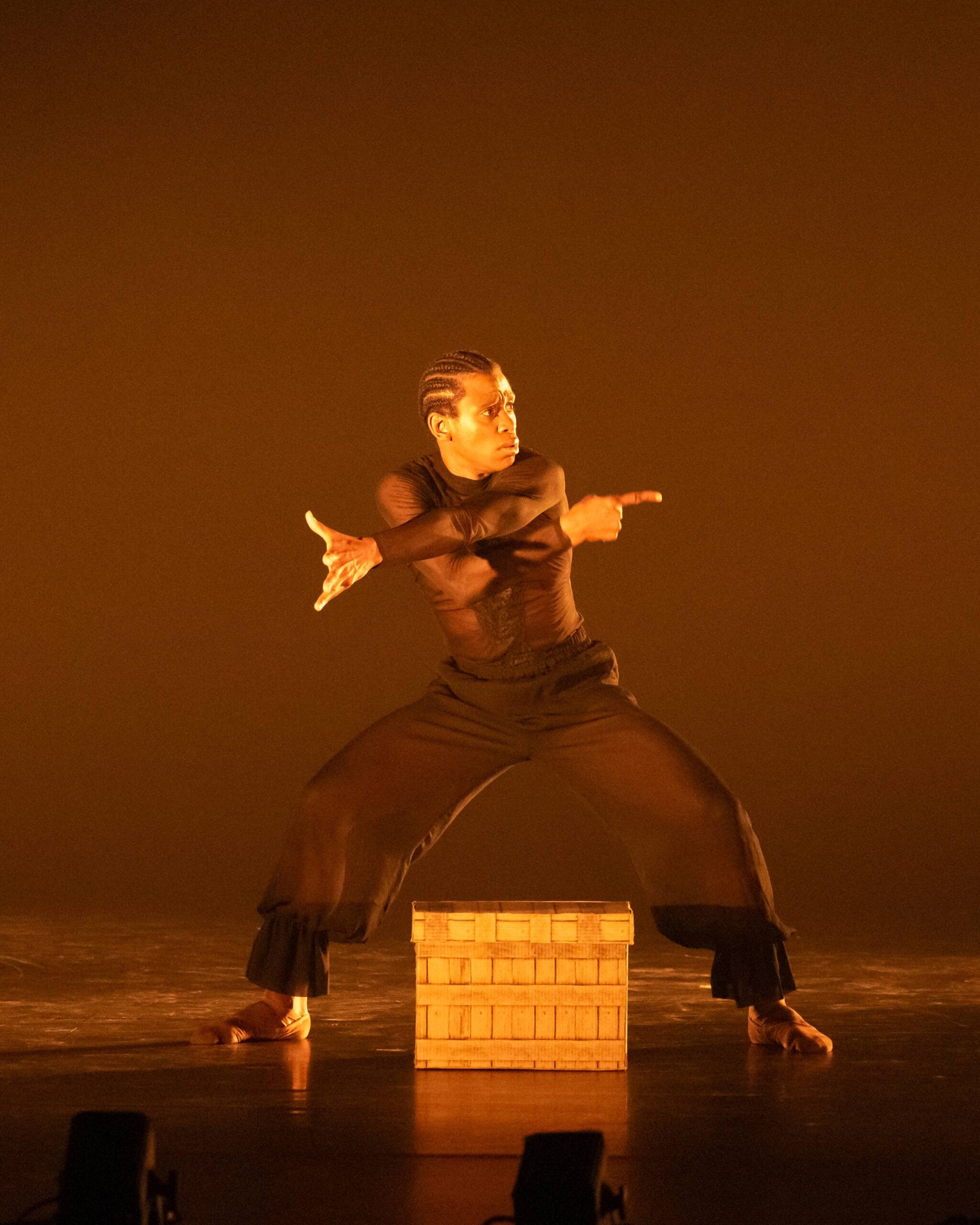 A dancer in black clothing performs a dramatic pose on stage in front of a wooden crate, against an orange-lit background.
