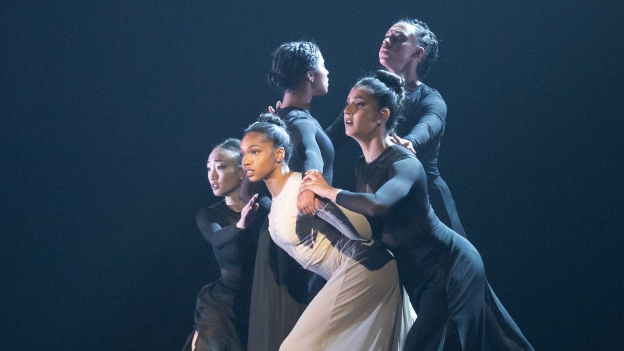 Five dancers in black and white costumes perform a contemporary dance on stage, standing closely together and holding each other in a dramatic pose under stage lighting.