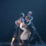 Five dancers in black and white costumes perform a contemporary dance on stage, standing closely together and holding each other in a dramatic pose under stage lighting.