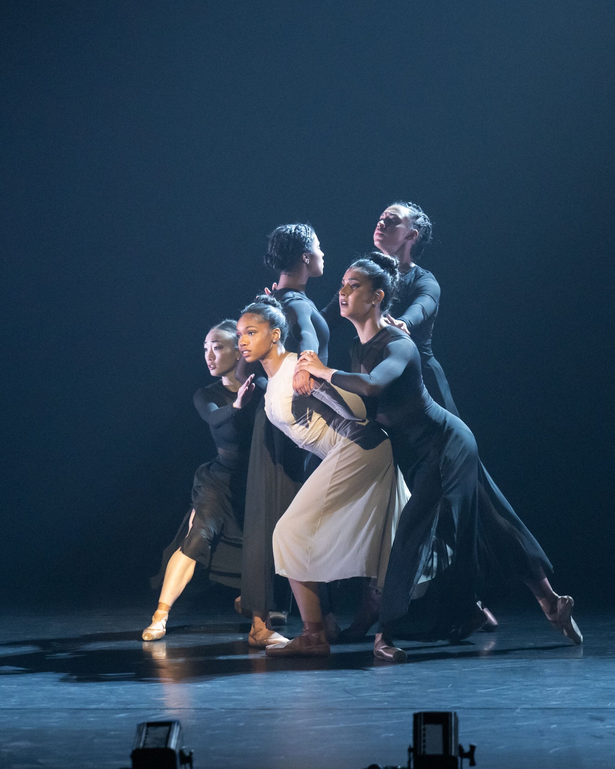 Five dancers in black and white costumes perform a contemporary dance on stage, standing closely together and holding each other in a dramatic pose under stage lighting.
