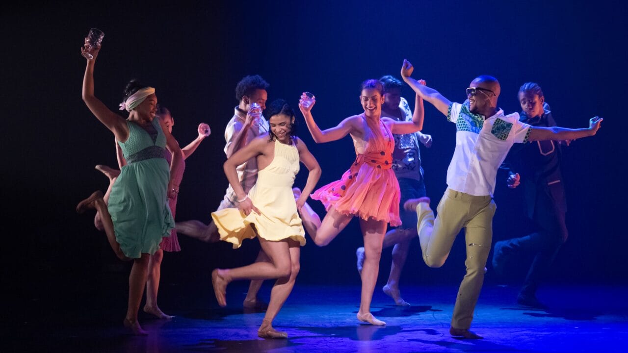 A group of barefoot dancers in colourful outfits perform energetically on stage under blue lighting.