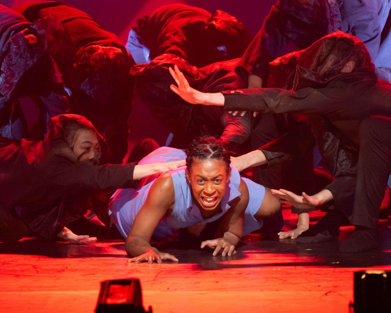 A performer in a blue outfit crawls on stage with an intense expression, surrounded by dancers in dark clothing reaching toward them under dramatic red lighting.