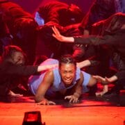 A performer in a blue outfit crawls on stage with an intense expression, surrounded by dancers in dark clothing reaching toward them under dramatic red lighting.