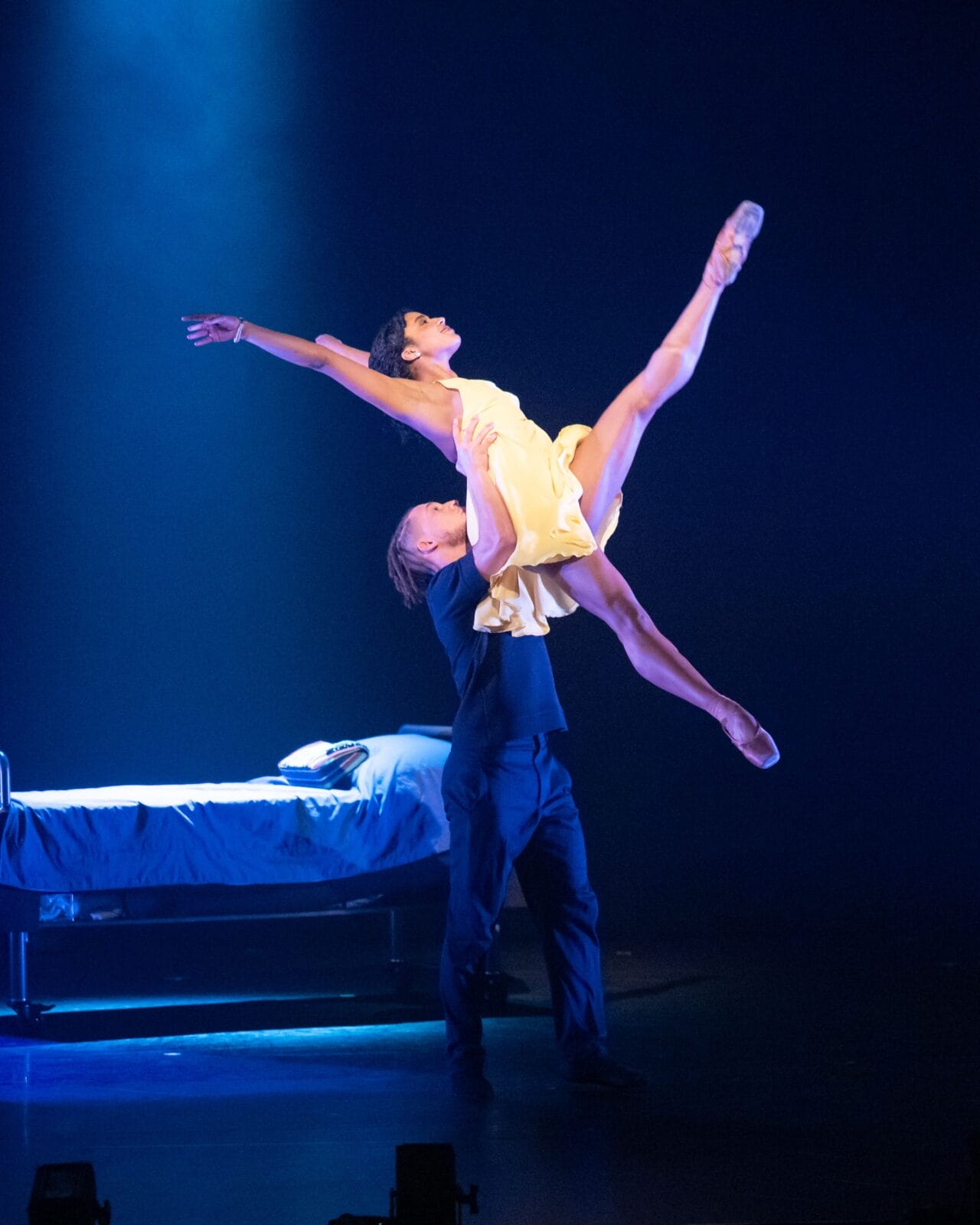 A male dancer lifts a female dancer in a yellow dress with one leg extended, performing on stage next to a bed under dramatic lighting.