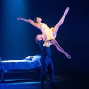 A male dancer lifts a female dancer in a yellow dress with one leg extended, performing on stage next to a bed under dramatic lighting.