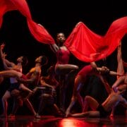 A group of dancers pose dramatically on stage, with one dancer in the center holding flowing red fabric above her head.
